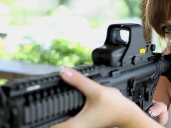 Woman looking through a holographic sight of an AR-15 rifle.