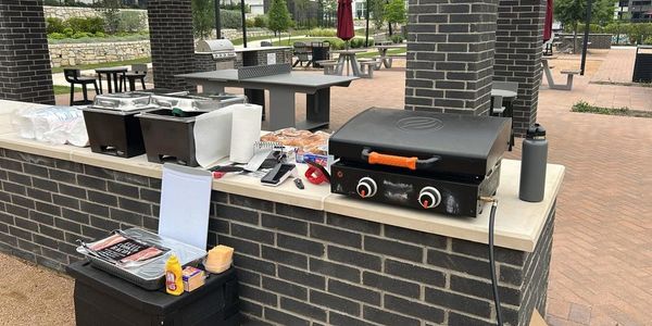 Outdoor barbecue setup with grill, buns, and condiments under a covered patio.
