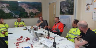 Workers in safety gear discuss at a meeting table with samples and documents.