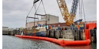 Crane lifting a boat from water near dock with orange containment boom.