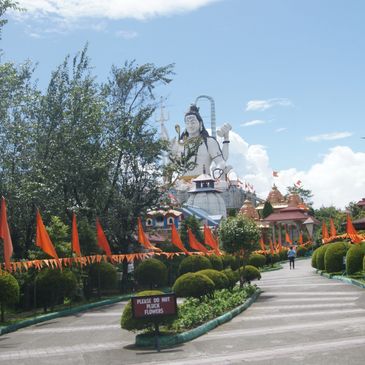 Pathway lined with orange flags leading to a large Shiva statue under a clear sky.