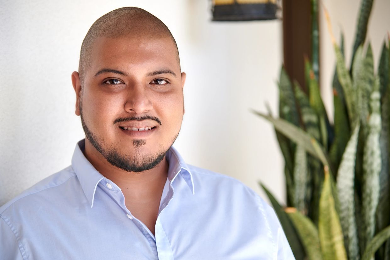 Smiling man in a light blue shirt standing indoors with plants in the background.