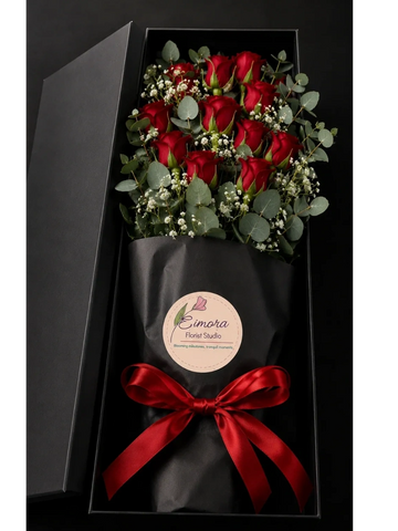 A bouquet of red roses with greenery in a black gift box tied with a red ribbon.