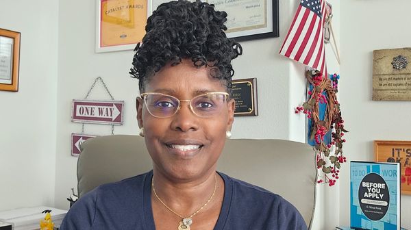 Smiling woman with curly hair and glasses sits in an office with awards and an American flag.