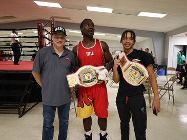 Two boxers proudly display championship belts with a man in an Army cap beside them.