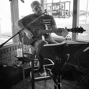 black and white photo of man sitting down playing acoustic guitar with a notebook in front of him