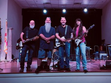 4 people  standing on stage. One man is holding a bass, the other man and women are holding guitars.