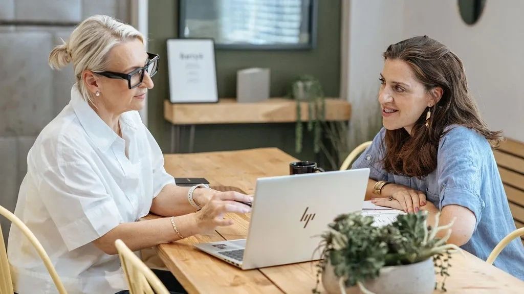 Two women sitting at a table with a laptop smiling and talking