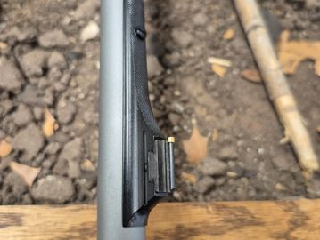 Close-up of a black firearm part on a wooden surface with dirt background.