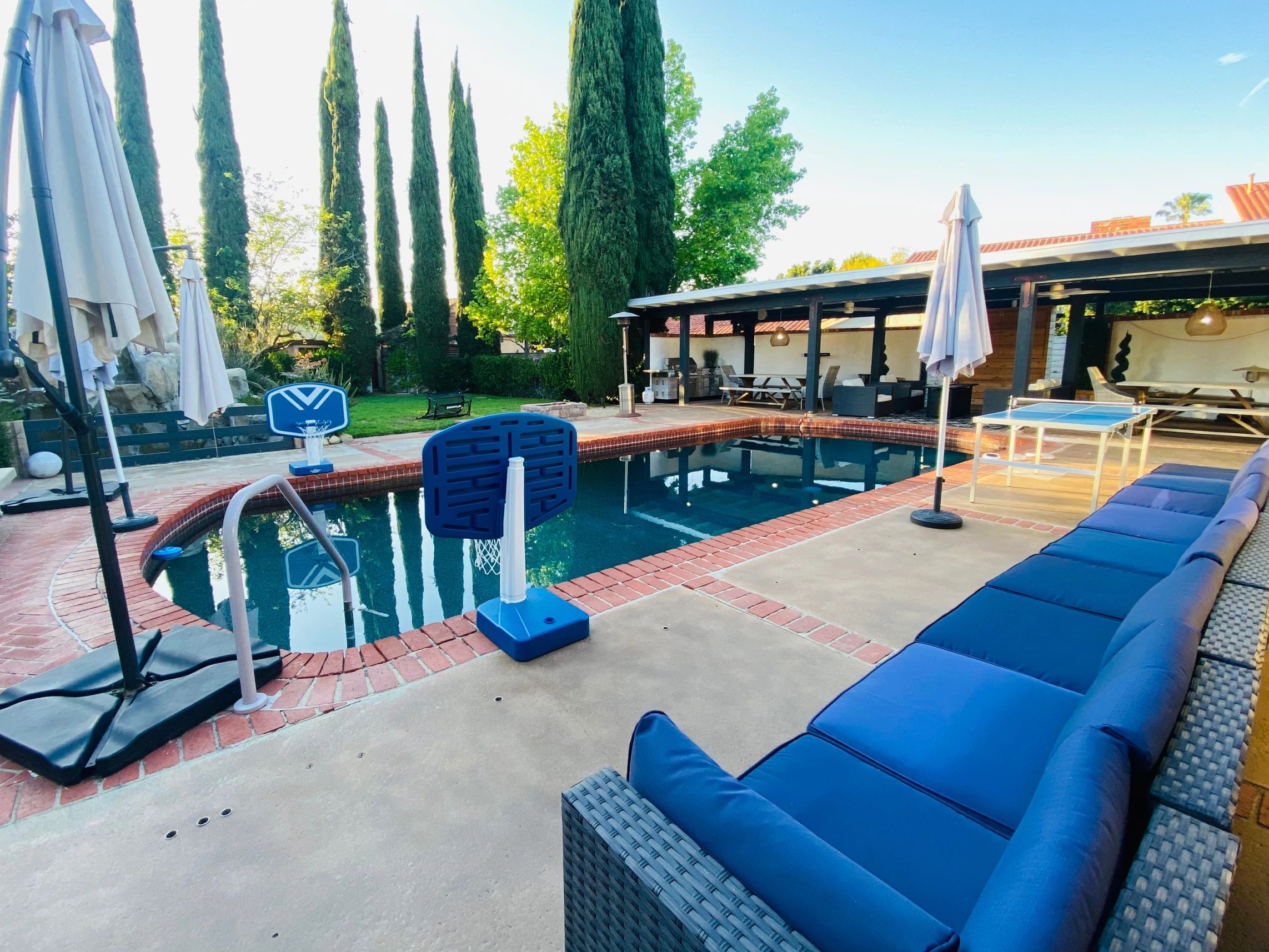 Outdoor pool area with lounge seating, mini basketball hoops, and tall trees in the background.