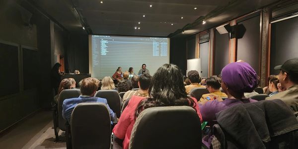 Audience watching a panel discussion in a dimly lit room with a large screen.