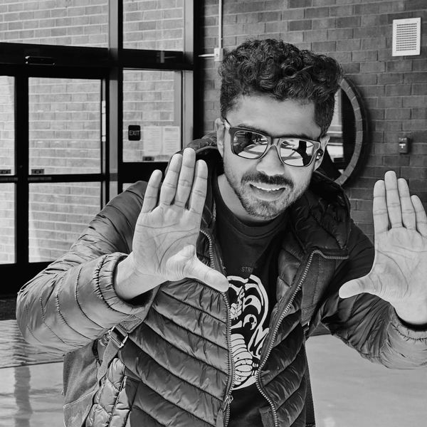 Young man in sunglasses and puffer jacket posing with hands up indoors.