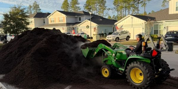 A man operates a green tractor to move a large pile of soil in a residential area.