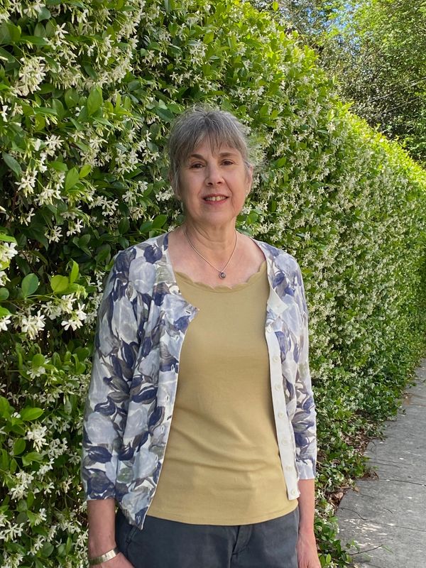 A woman standing in front of a lush green hedge with white flowers.