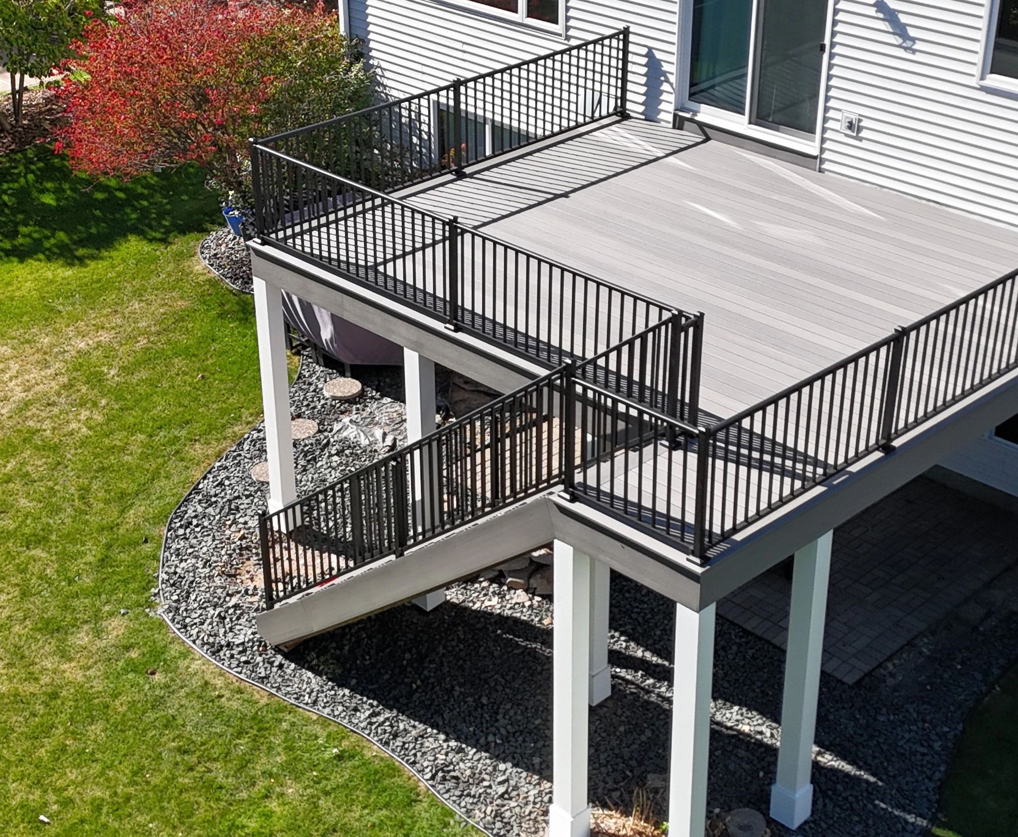 Elevated deck with black railing and stairs leading to a backyard with green grass and a red tree.