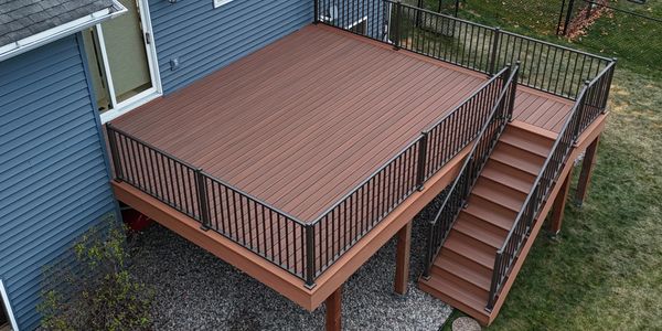 A spacious brown wooden deck with black railing attached to a blue house.