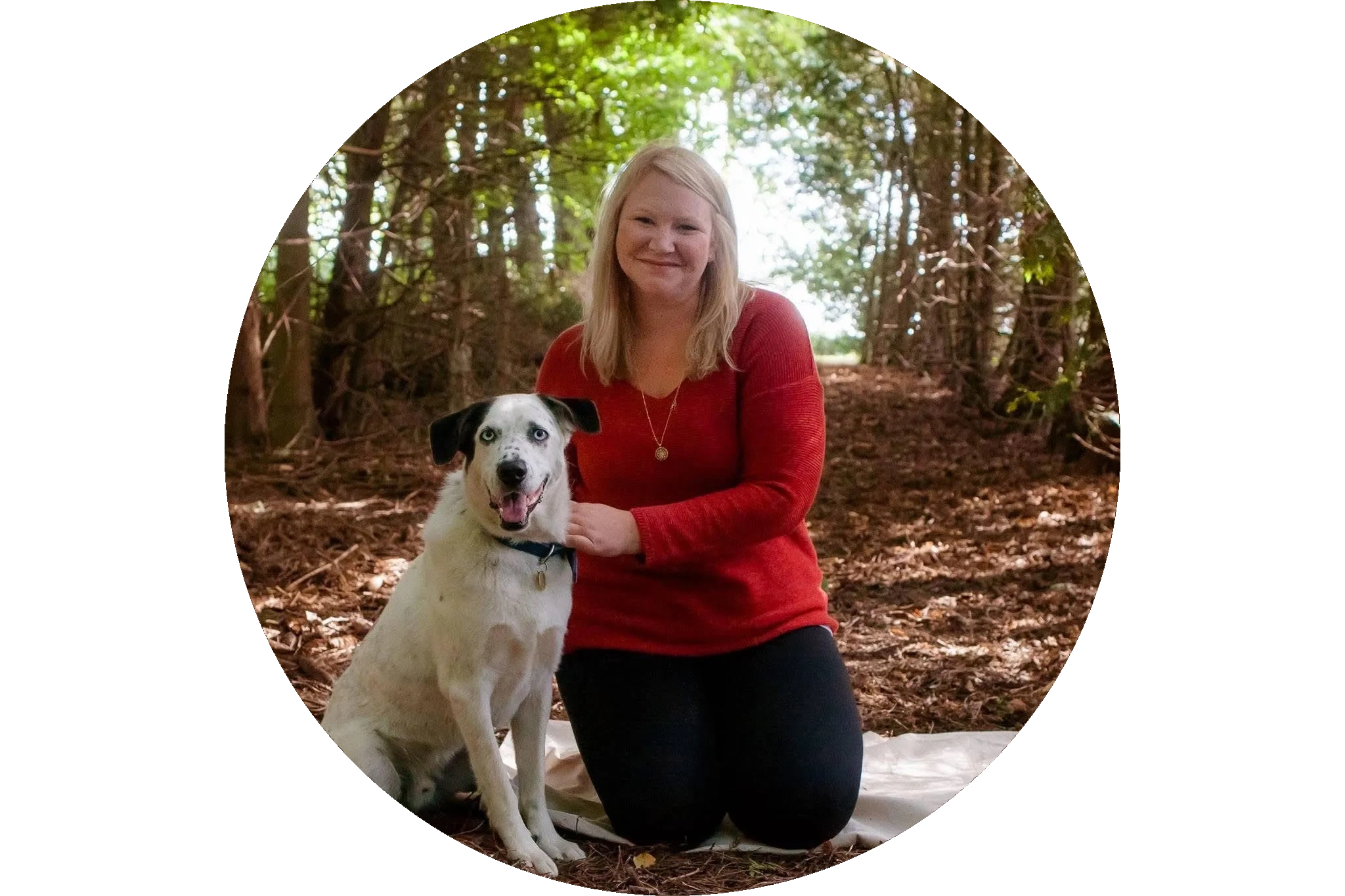 Woman in a red sweater kneeling beside a white dog in a wooded area.