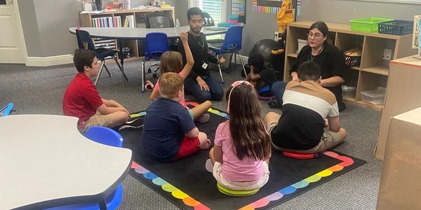 A diverse group of children and two adults sit in a classroom circle for a discussion.