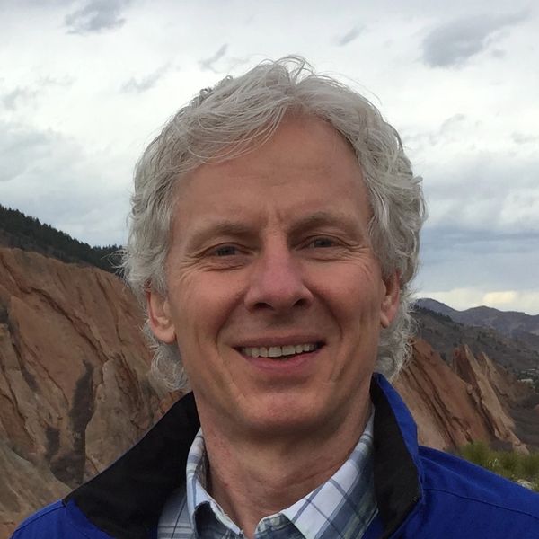 Smiling man with gray hair outdoors near rocky hills under a cloudy sky.