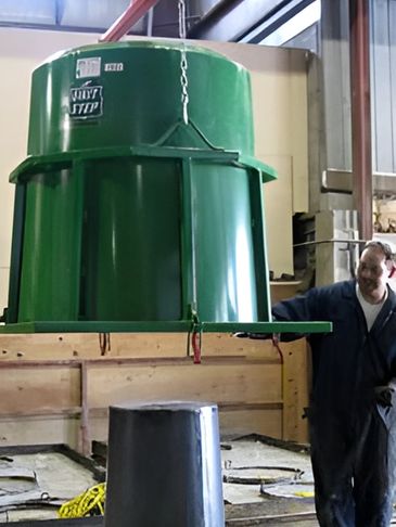 A worker maneuvers a large green cylindrical container suspended from a crane in a factory setting.