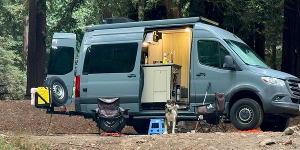 Camper van setup in the woods with chairs and a dog outside.
