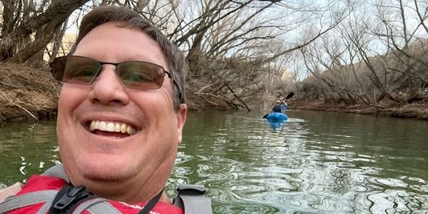 Man smiling on a calm river with another person kayaking behind.