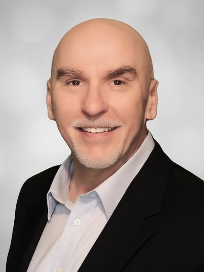 Smiling bald man in a black blazer and light blue shirt against a gray background.