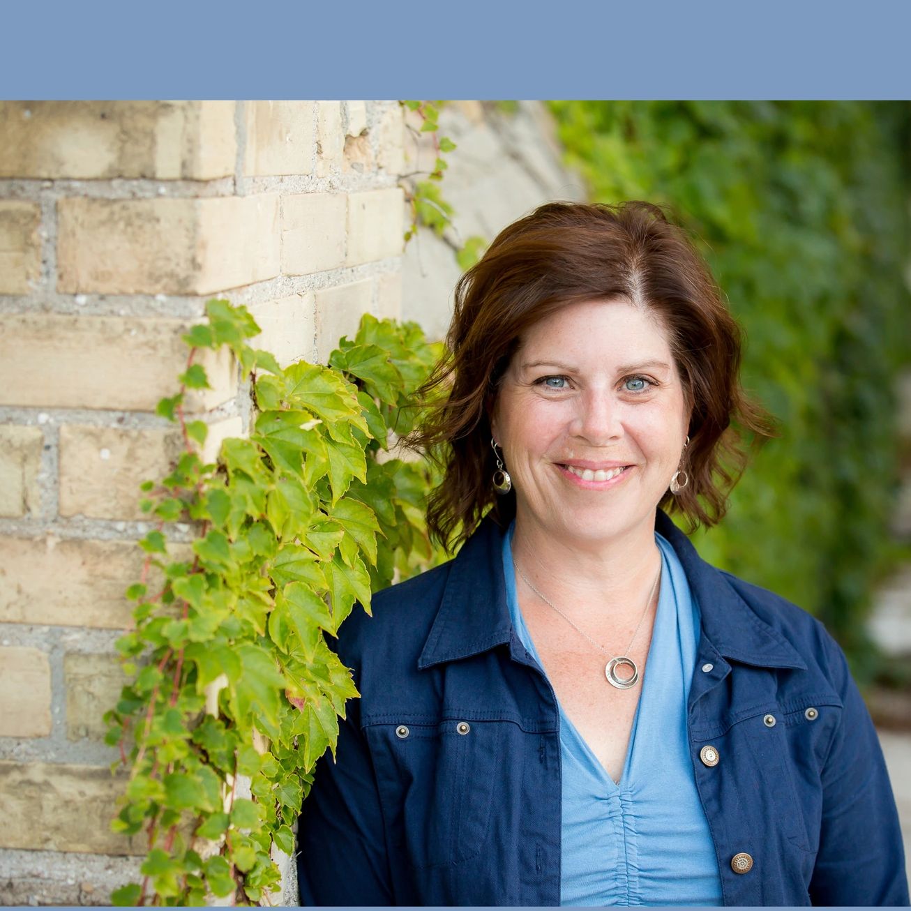 Bethany Mazereeuw smiling in blue jacket stands by ivy-covered brick wall.