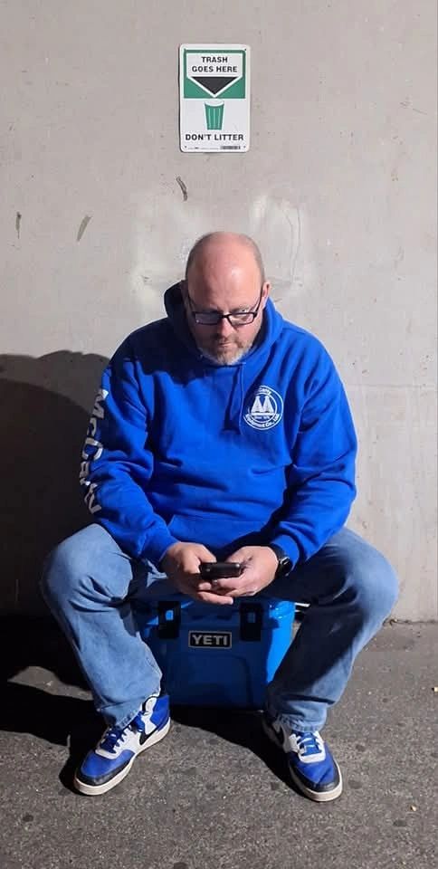 Man in blue hoodie sitting on a Yeti cooler under a trash sign.