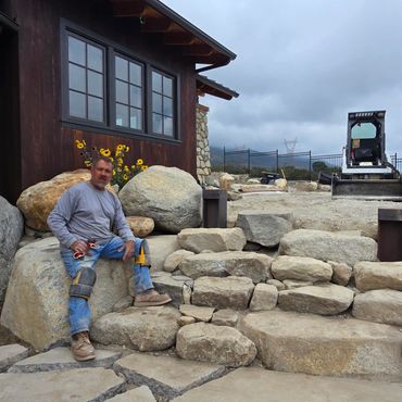 Man sitting on large stone steps outside a rustic building under cloudy sky.