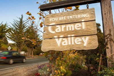 Wooden sign welcoming visitors to Carmel Valley beside a road.