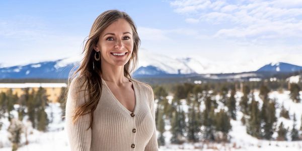 Smiling woman in beige sweater with snowy mountain background.