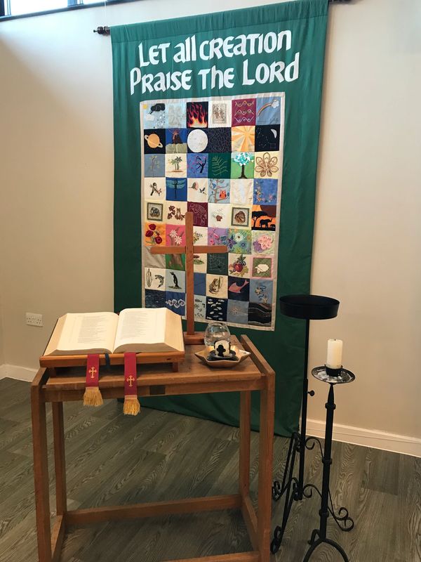 An open bible, wooden cross and candle in front of a patchwork tapestry. 