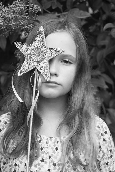 Girl holding a star-shaped wand covering one eye, surrounded by foliage.