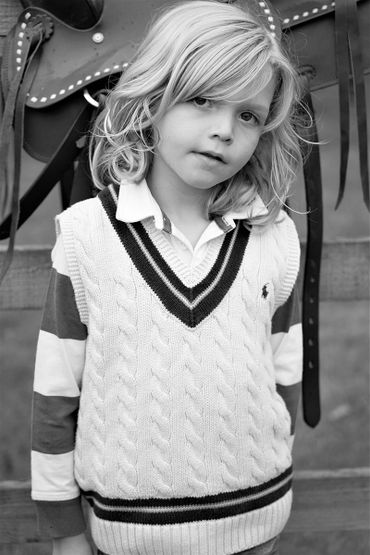 Blond child in a cable-knit sweater vest posing in front of a saddle.