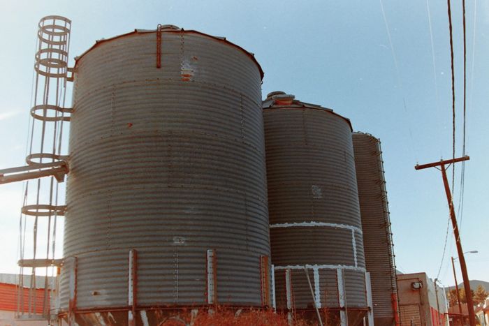 Three metal grain silos, with a clear blue sky in the background and powerlines on the right.