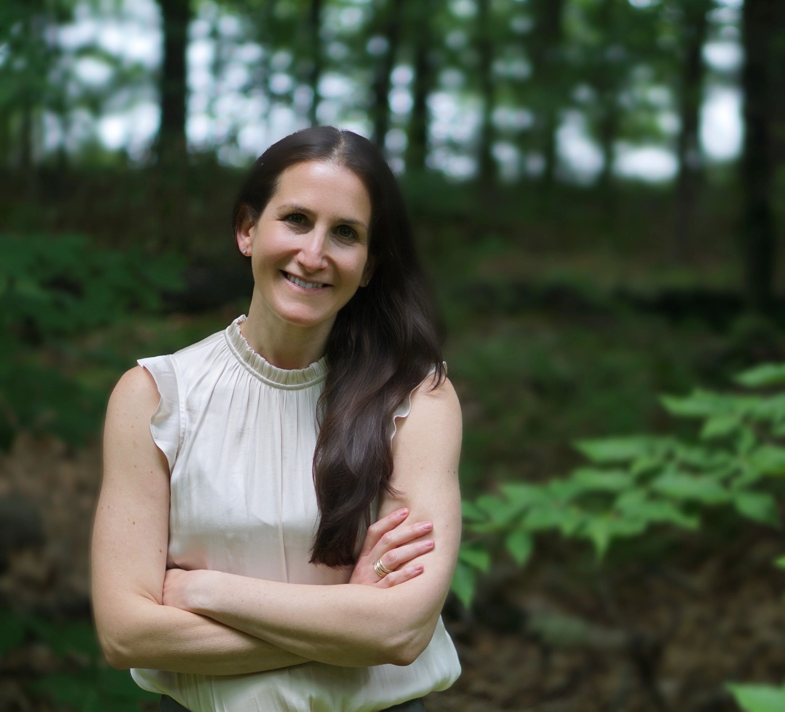 A picture of Julie standing in the woods, smiling with her arms crossed in a cream colored shirt.