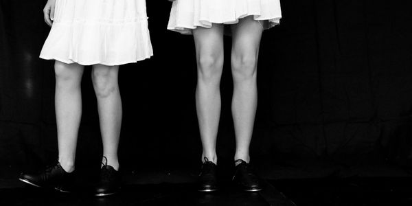 Two girls in white dresses and black shoes against a dark backdrop.