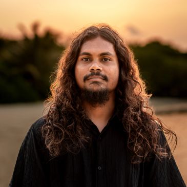 Man with long curly hair and beard at sunset outdoors.