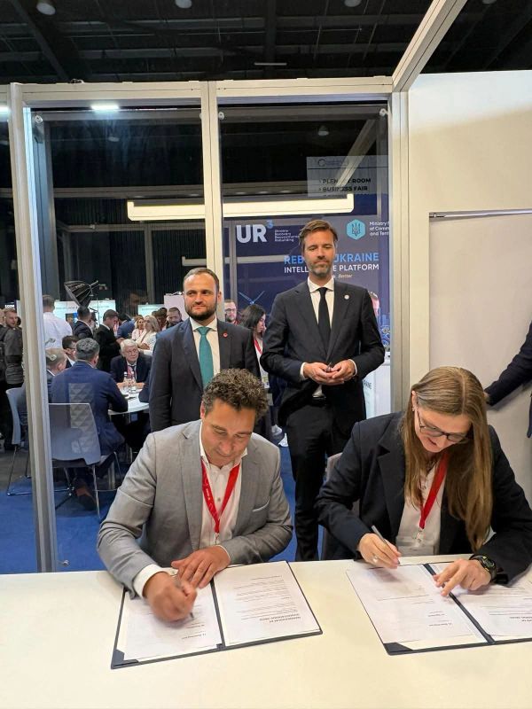 Two individuals signing documents at a formal event with observers behind them.