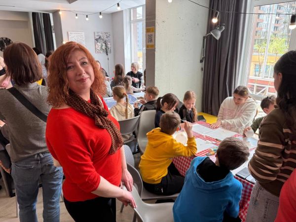 Woman in red leading a classroom with children at tables.