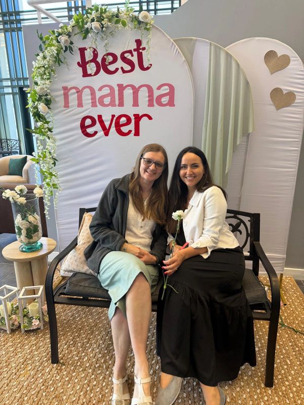 Two women sitting on a bench with a "Best mama ever" sign behind them.