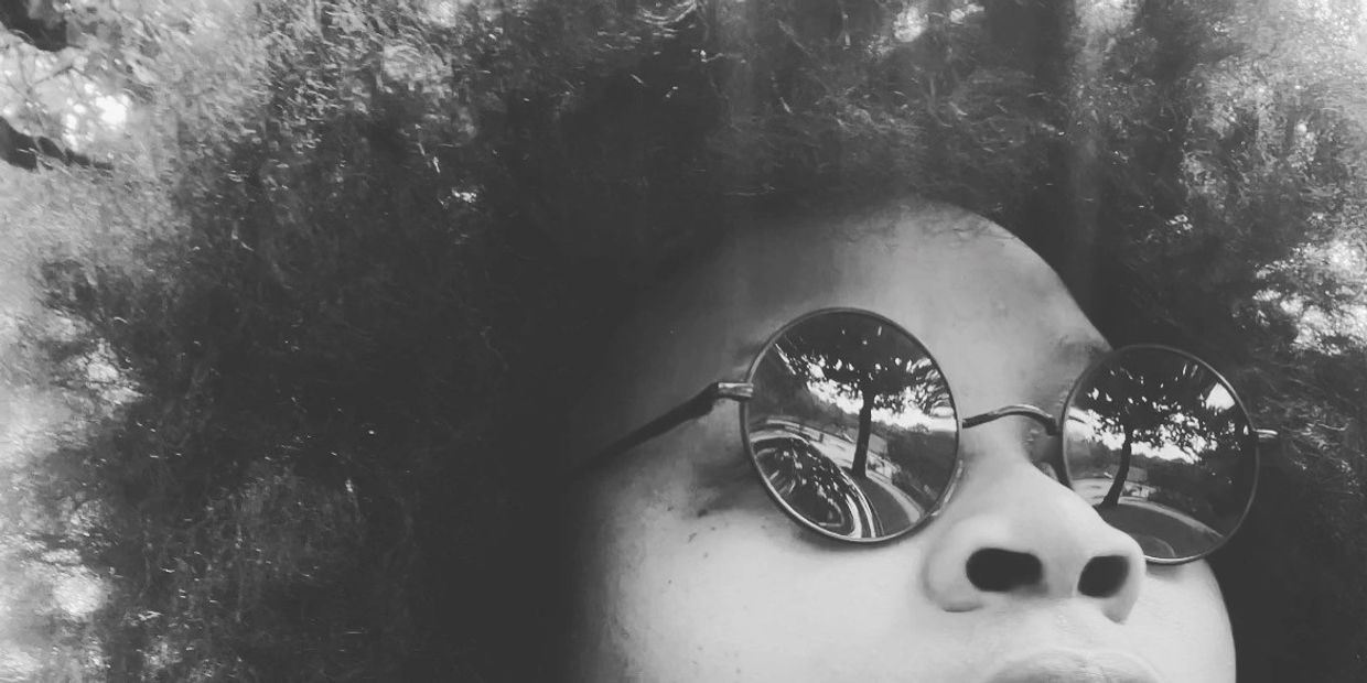Close-up of a person with round sunglasses reflecting trees and a path.