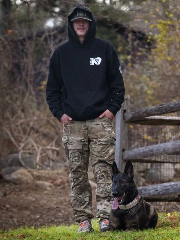 A young man in camo pants stands with a black dog sitting beside him outdoors.