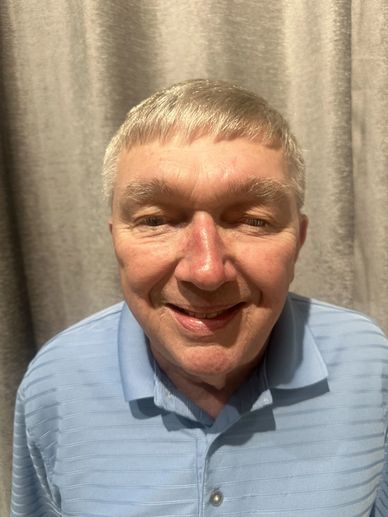 Smiling elderly man in a light blue polo shirt against a gray curtain background.