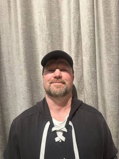Man in a black hoodie and cap standing against a gray curtain.