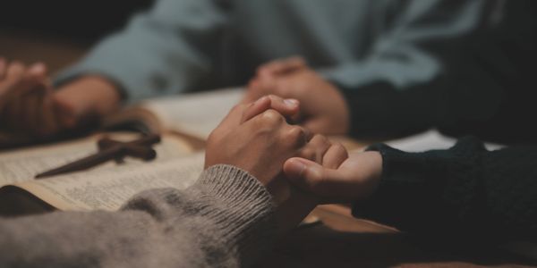 People holding hands over an open Bible with a wooden cross.