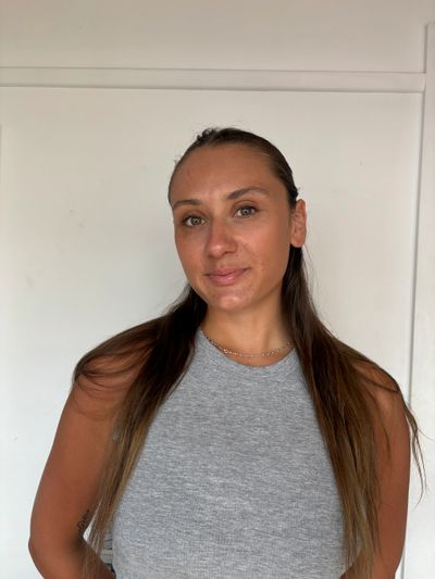 A woman with long brown hair wearing a gray sleeveless top, standing against a white wall.