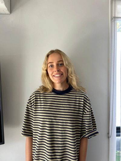 Young woman smiling, wearing a striped shirt, standing indoors against a white wall.
