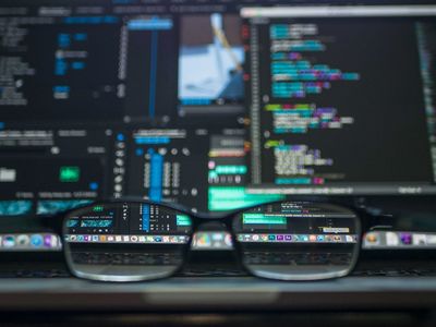 A pair of reading glasses on a table in front of multiple computer screens.